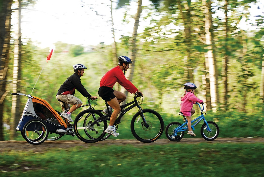 Eine Familie fährt Fahrrad auf einem Waldweg. Zwei Erwachsene auf Mountainbikes und ein Kind auf einem kleinen blauen Fahrrad, alle tragen Helme. Ein Kinderanhänger ist an einem der Erwachsenenräder befestigt.